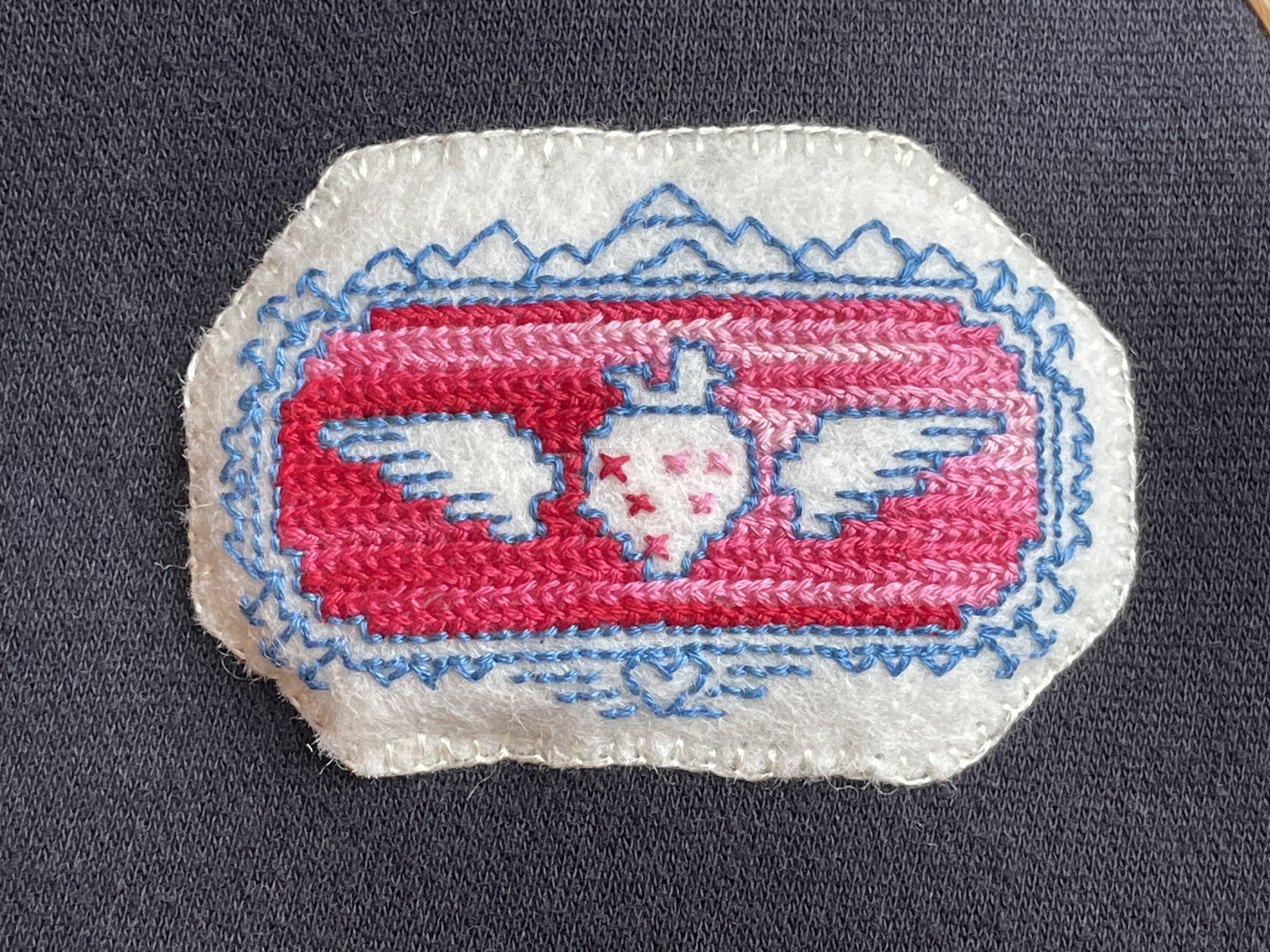 A felt patch featuring a voided-work winged strawberry, with a decorative border of mountains, arrows, spikes, and a crystal heart.