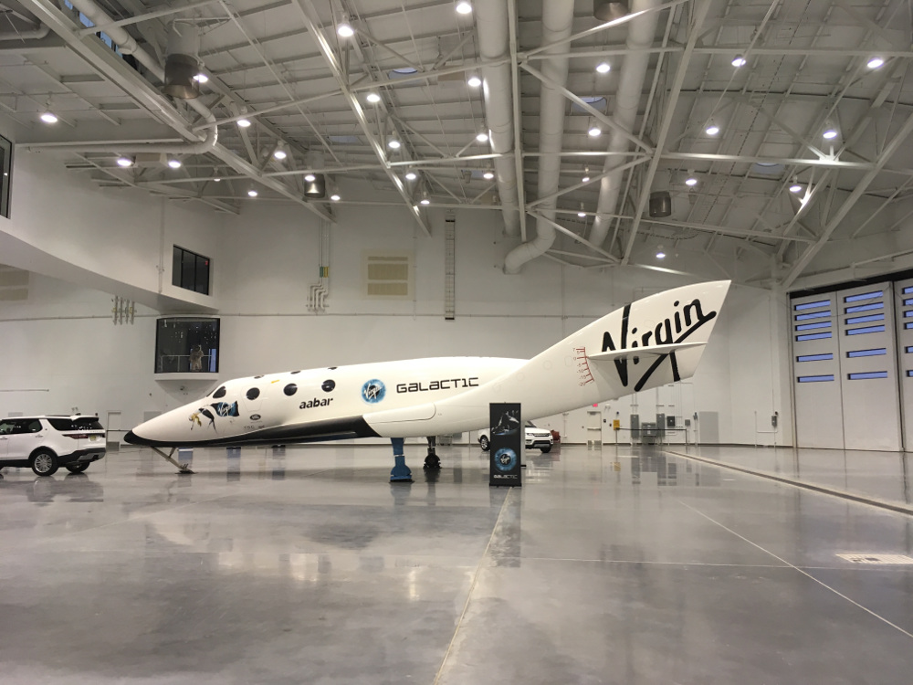 A "space plane"-style spacecraft inside a large hangar.  The ship features Virgin and Virgin Galactic logo decals, as well as the word "aabar".